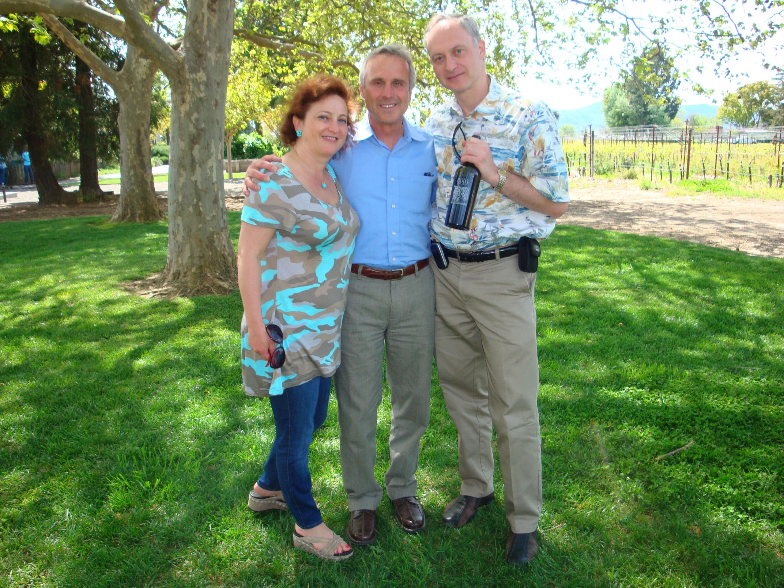 3 people smiling holding Biale wine bottle in grass with trees and vineyards in background