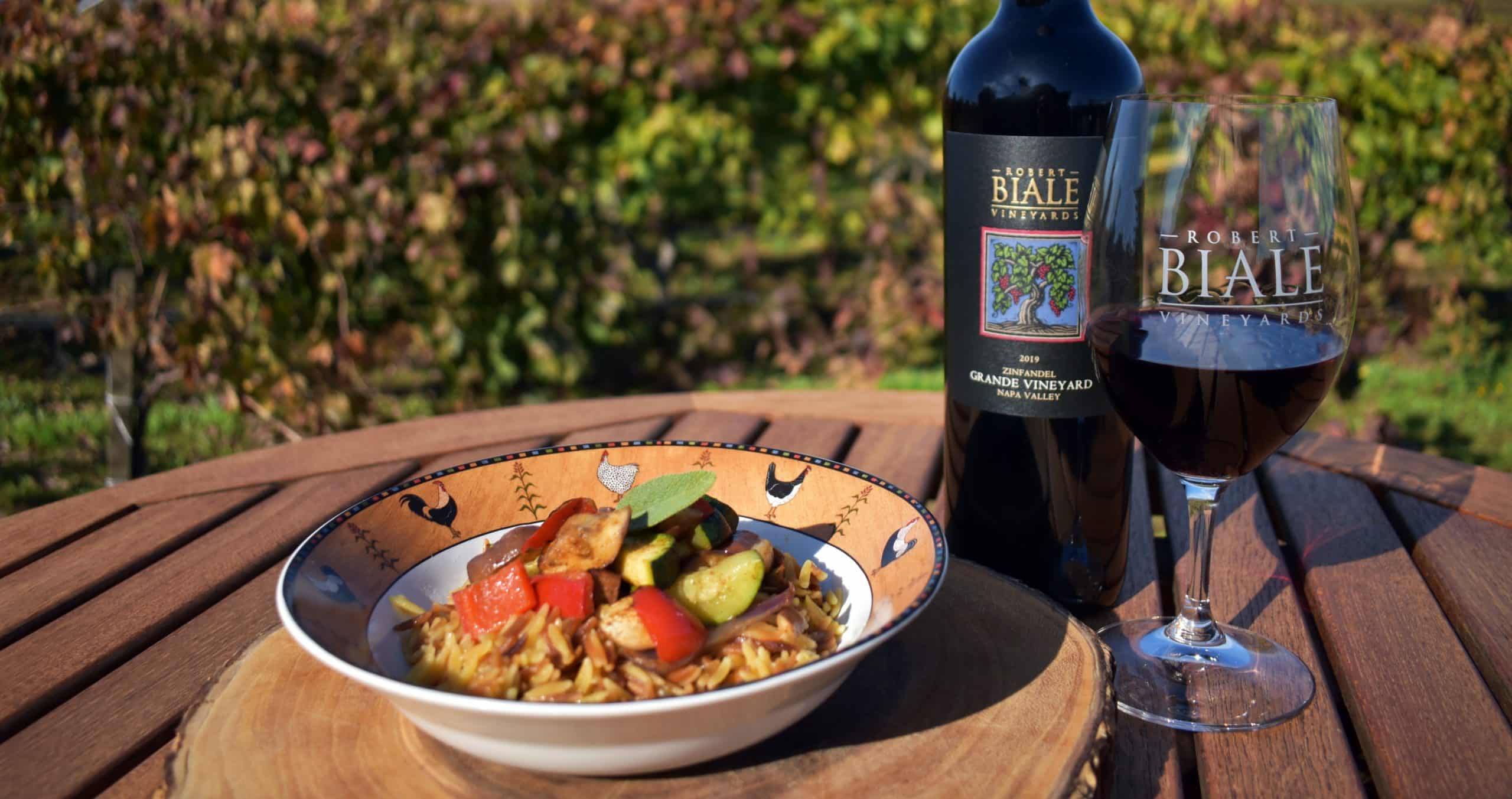 dish filled with orzo and vegetables on wooden table accompanied by a bottle of Biale wine and filled wine glass with Biale logo