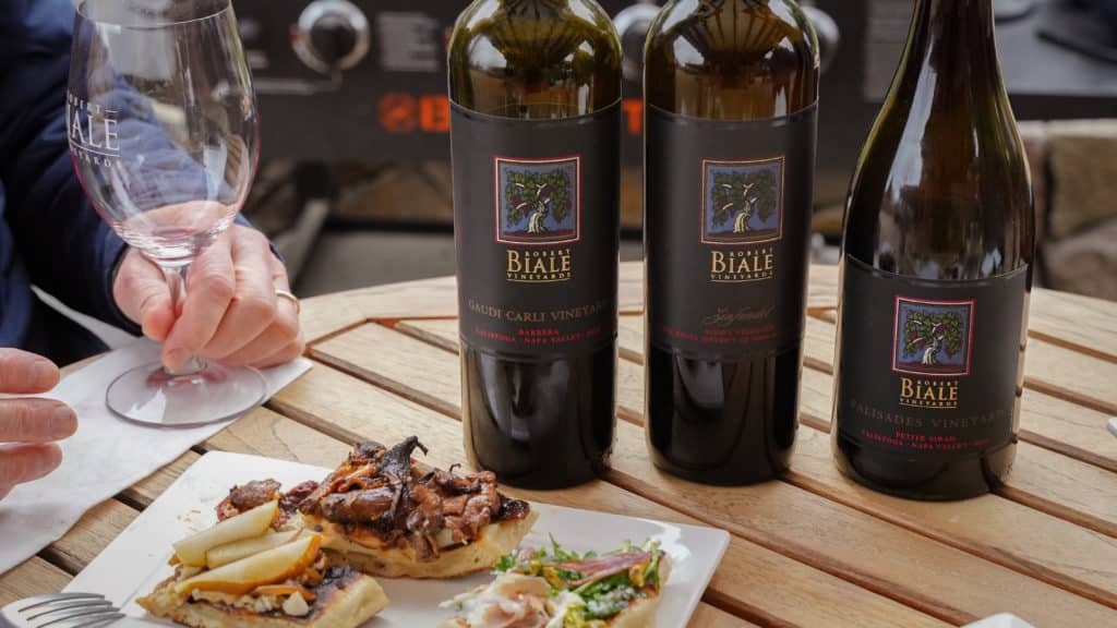 three bottles of Biale wines on table with plate of topped flatbreads, hand holding Biale logo wine glass on left side of table
