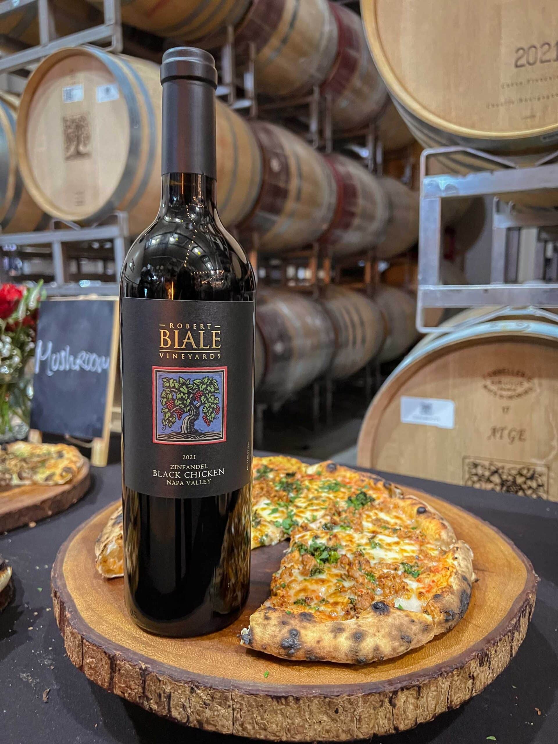 a bottle of Biale Black Chicken Zinfandel beside a pizza in the Biale cellar with barrels in the background